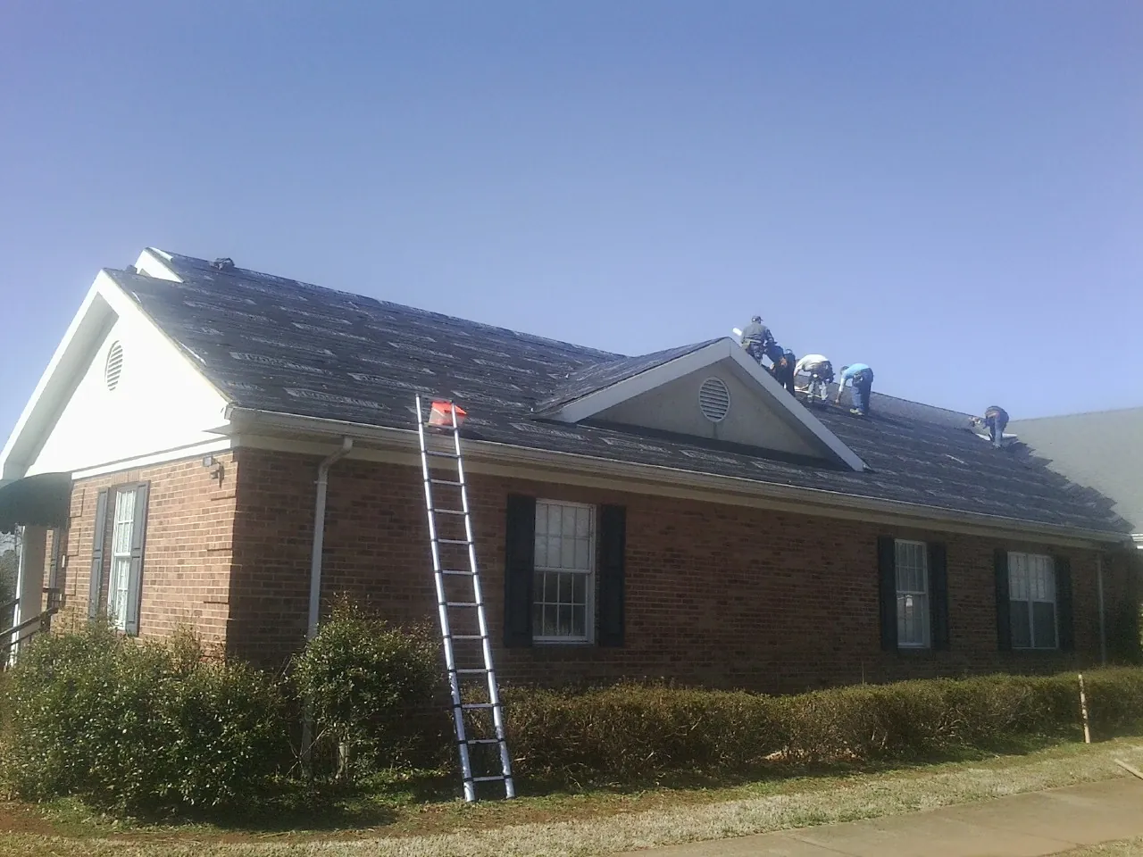 Crew tearing shingles off a brick ranch roof with a ladder set — Emergency Roof Repair work by our crew in Clayton